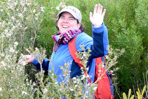 Amelia, stood amongst plants smiling and waving to the camera. She is wearing a beige baseball cap, a pink scarf and carrying a red rucksack